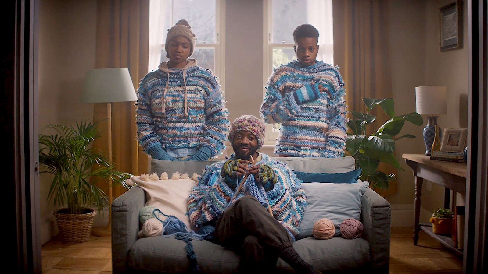 A father sitting on a sofa knitting with two teenagers standing behind him in hand knitted jumpers