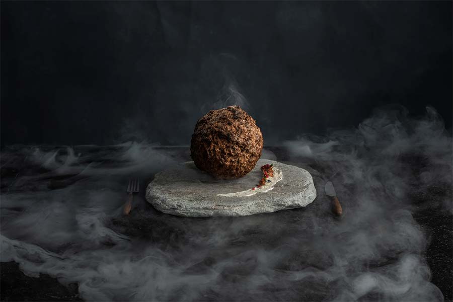 A large meatball made of mammoth meat on a stone platter in a dark studio set with mist swirling around it